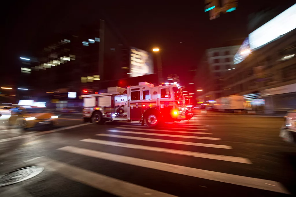 Fire engine with lights on in intersection at night on busy city street.